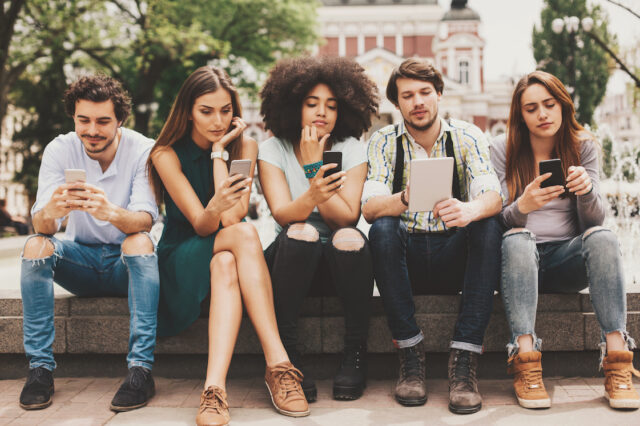 Group of mixed race people looking at mobile phones and tablets outdoors at urban setting
