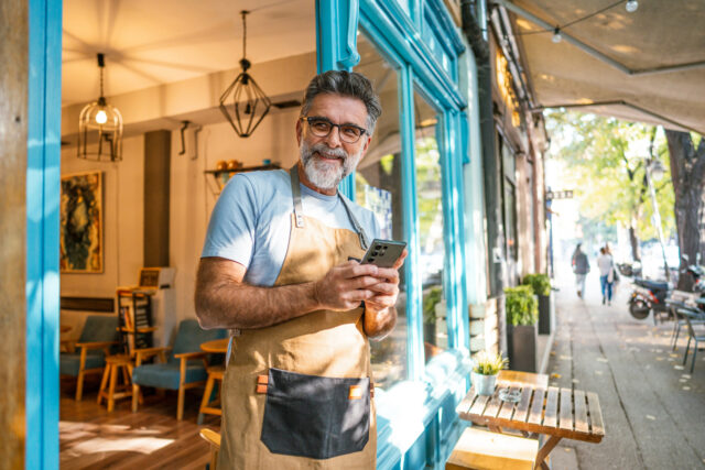 Mature man owner with an apron using phone in front of the café